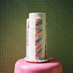 A novelty toilet paper holder shaped like a pink cake topper, holding three rolls of white toilet paper printed with colorful diagonal stripes, set against a green tiled wall background.