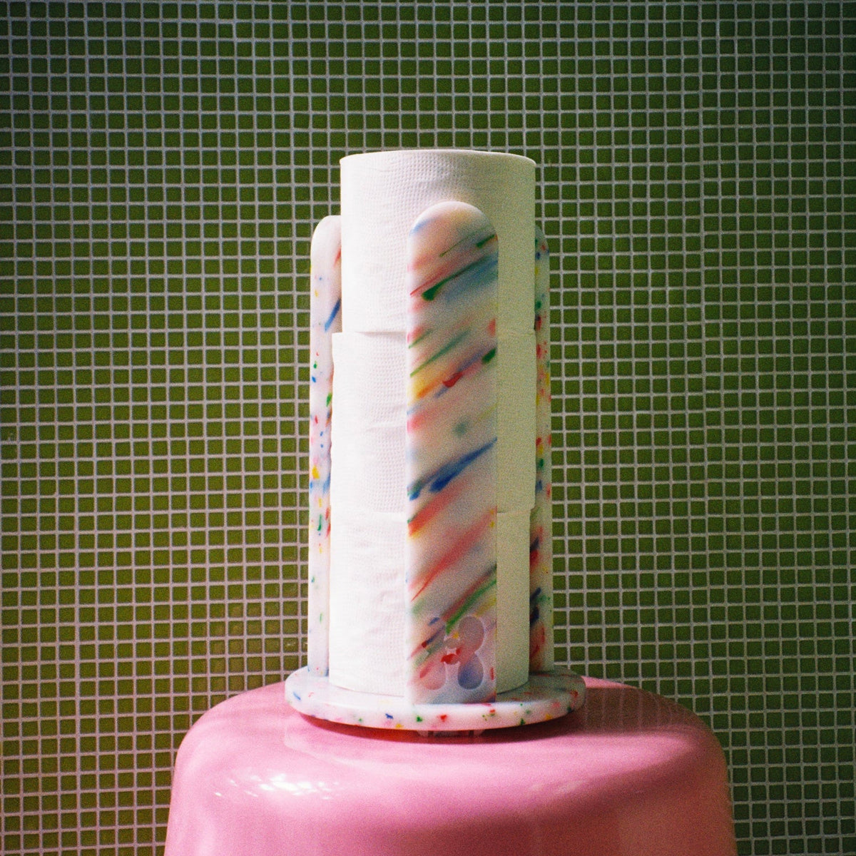 A novelty toilet paper holder shaped like a pink cake topper, holding three rolls of white toilet paper printed with colorful diagonal stripes, set against a green tiled wall background.