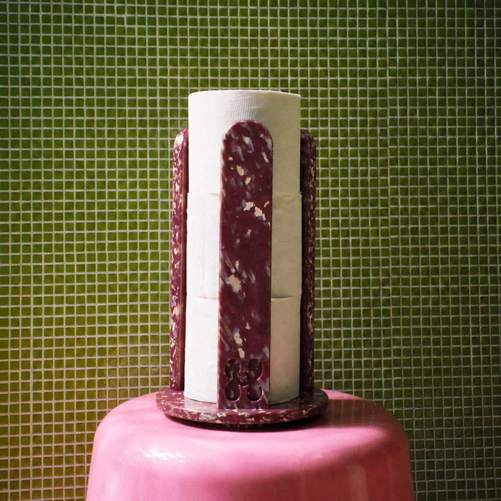 A decorative toilet roll holder with white and burgundy marbled vertical panels, displayed on a pink pedestal against a green tiled background.