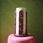 A decorative toilet roll holder with white and burgundy marbled vertical panels, displayed on a pink pedestal against a green tiled background.